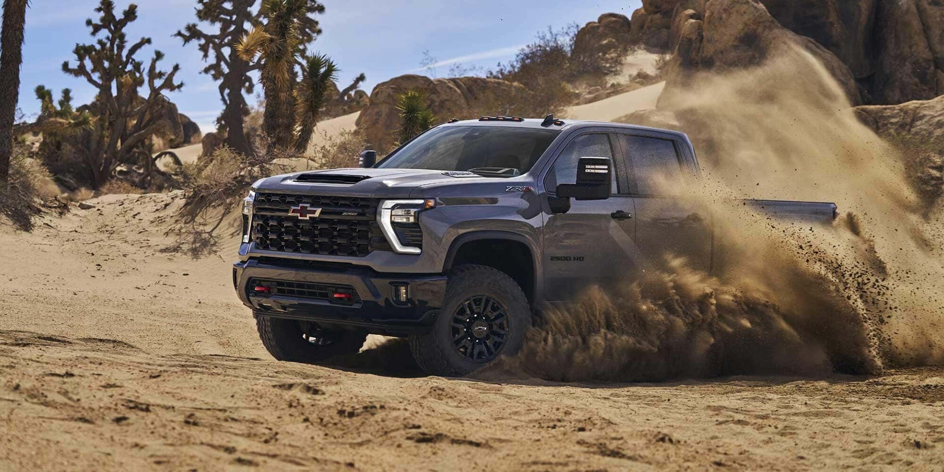 A gray Chevrolet pickup truck drives through a sandy desert landscape with clouds of dust swirling around. In the background, there are rocky formations and Joshua trees under a clear blue sky.