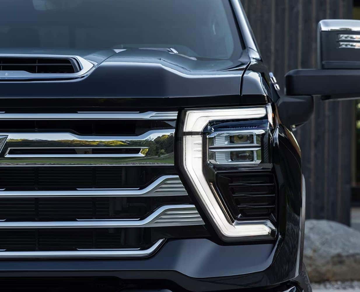 Close-up of the front of a black truck, showing a modern and sleek headlight design with LED accents. The grille features horizontal chrome bars, giving the vehicle a robust and stylish appearance. Part of the side mirror is visible.