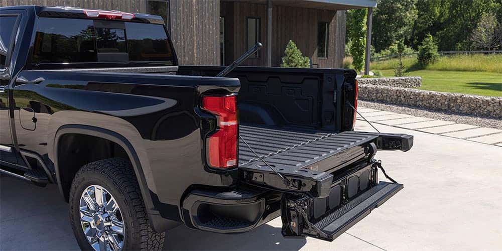 A black pickup truck parked on a driveway with its tailgate and step partially open. The truck has chrome wheels, and theres a modern wooden building with a stone wall and greenery in the background.