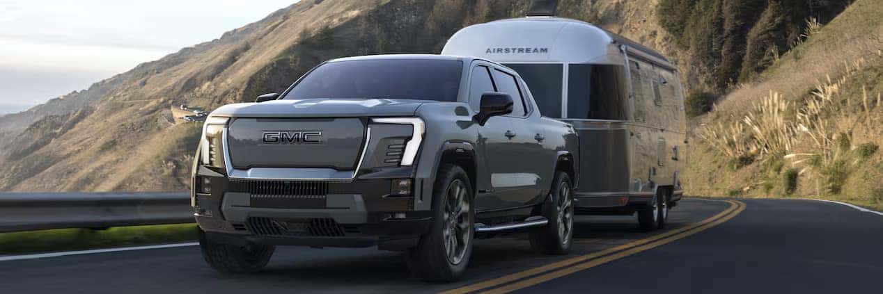 A silver GMC truck is towing an Airstream trailer on a winding road with scenic mountainous terrain in the background. The sky is clear, and the road curves around a hillside, suggesting a journey in a picturesque location.