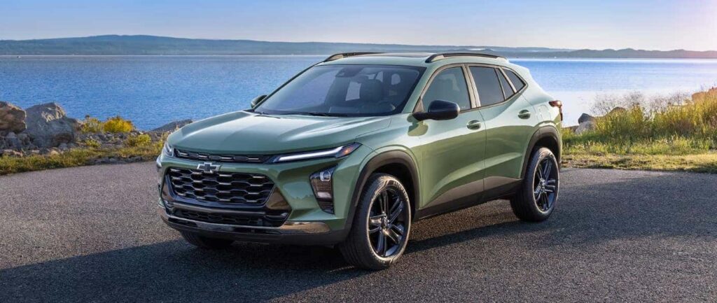 A green SUV is parked on a paved surface near a calm body of water. The car features a prominent grille and sleek design. In the background, hills are visible under a clear blue sky.