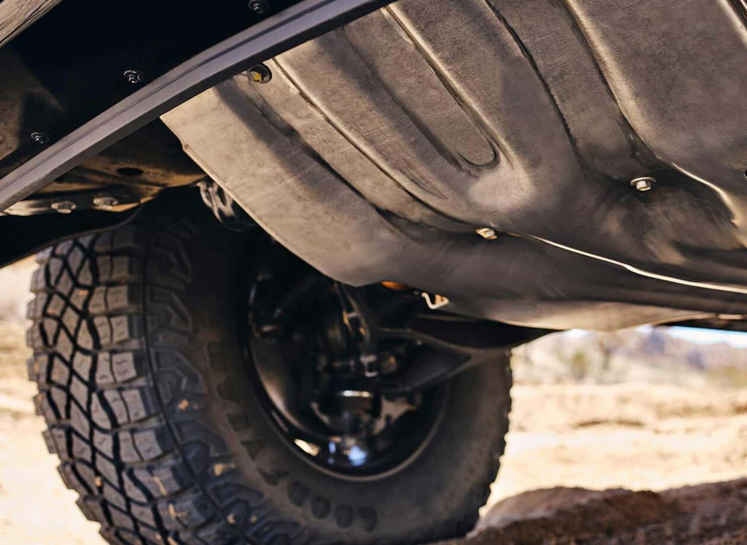 Close-up view of a vehicles undercarriage, showcasing a sturdy metal skid plate and large off-road tire with rugged tread. The image captures the vehicle outdoors, with a hint of a dusty landscape in the background.