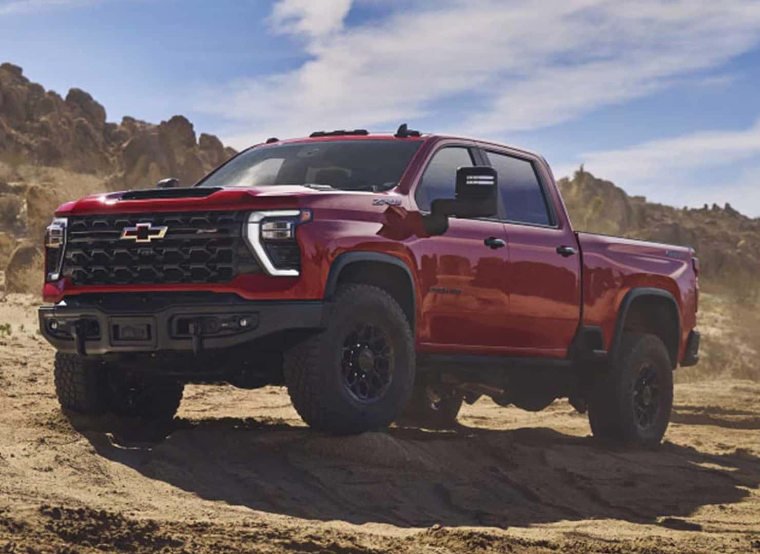 A red Chevrolet pickup truck parked on a sandy terrain with rugged hills in the background, under a clear blue sky. The truck features a modern design with black accents and all-terrain tires.