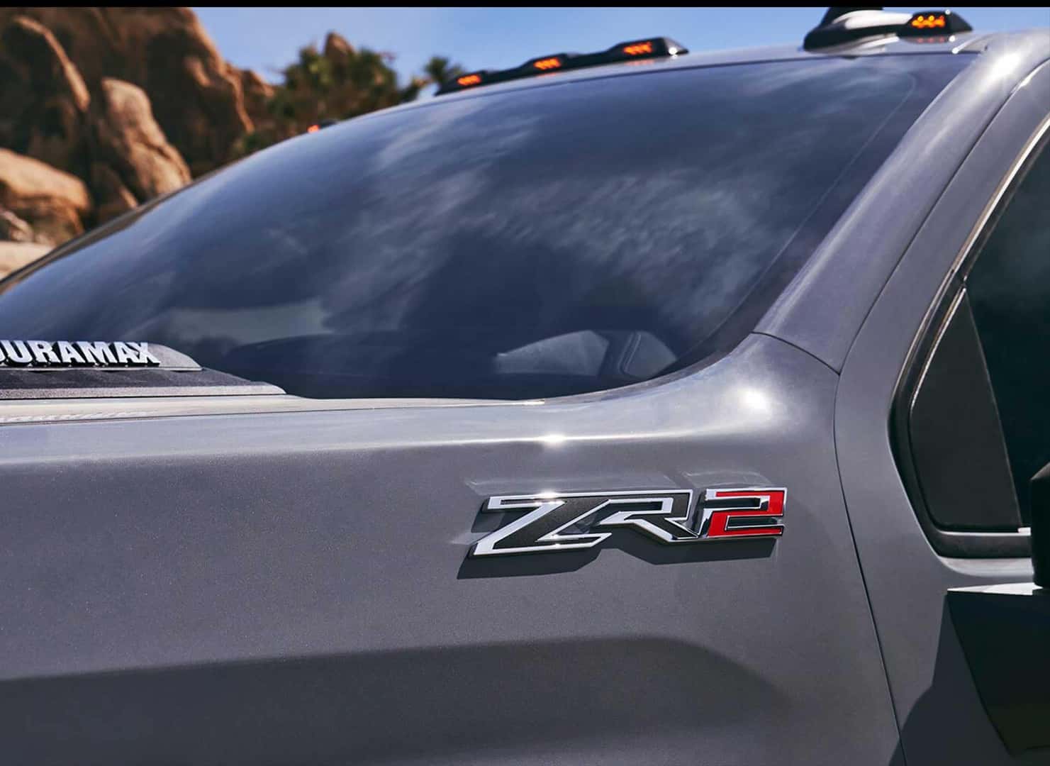 Close-up of a silver truck showing the ZR2 badge near the side mirror and Duramax label on the hood. The windshield is slightly tinted and the background features rocks and greenery under a clear sky.