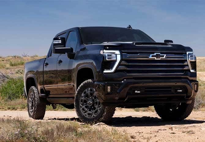A black pickup truck parked on a dirt road in a desert landscape. The vehicle is modern with prominent front grille and tires suited for rough terrain. The sky is clear and blue.