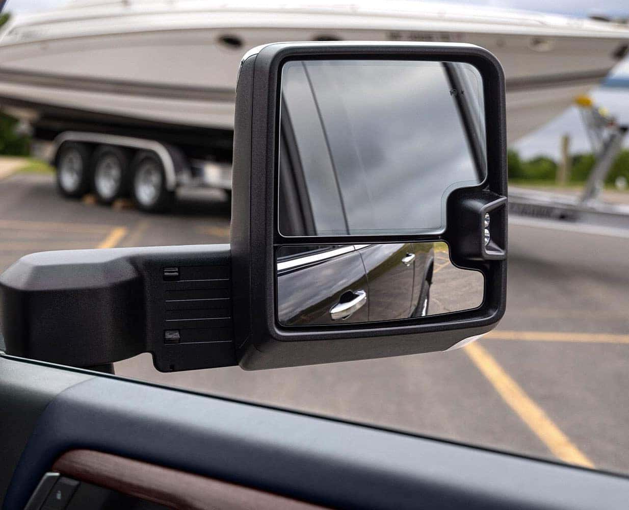 Close-up of a vehicles side mirror reflecting part of the car and an attached boat on a trailer. The mirror captures the boats white hull and several wheels of the trailer, with a parking lot and trees in the background.