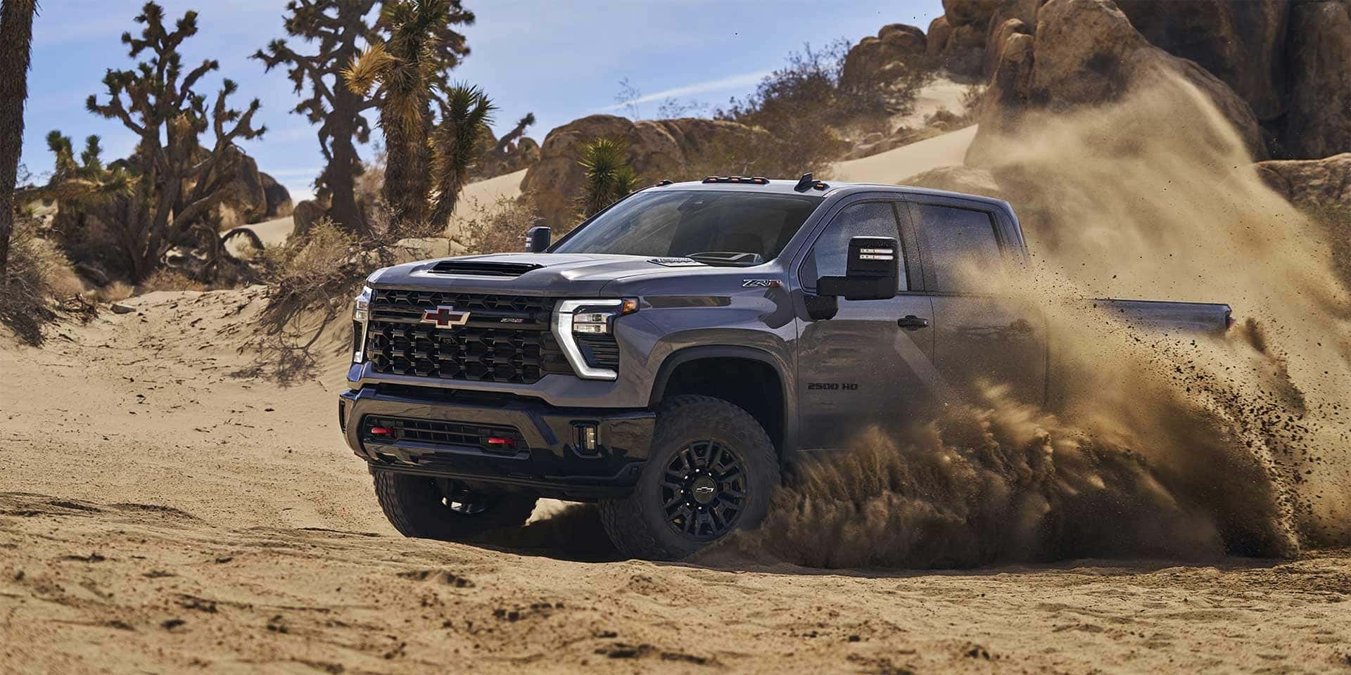 A black pickup truck drives through a sandy desert area, kicking up dust. The landscape features large rocks and Joshua trees under a clear blue sky. The truck has rugged off-road tires and a prominent grille.