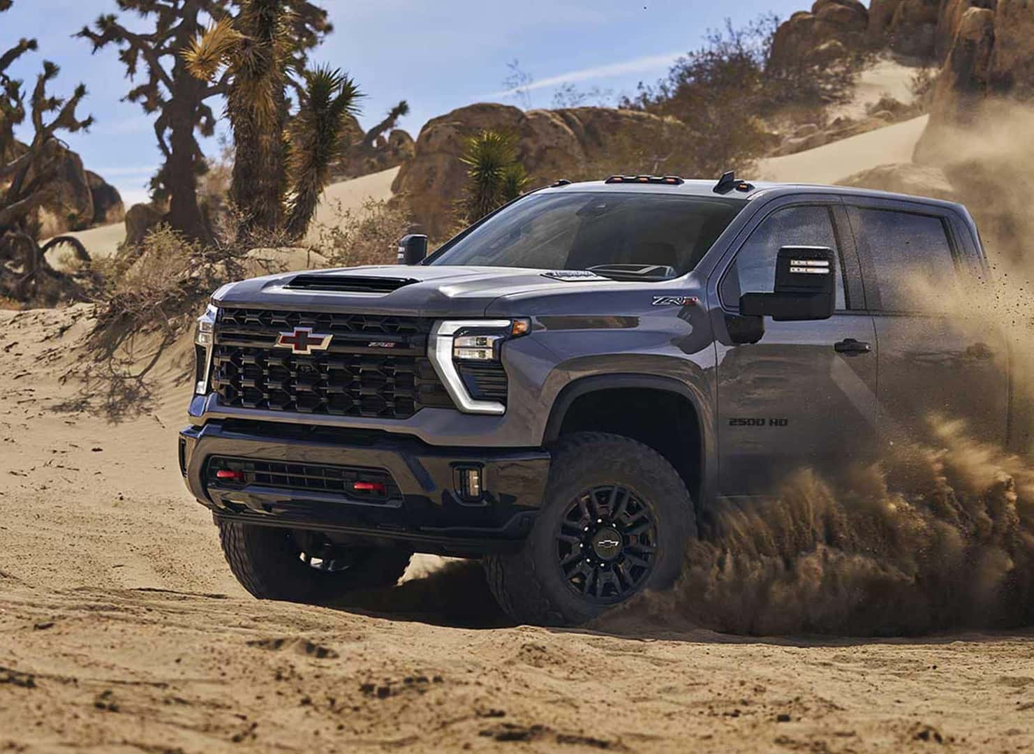 A rugged Chevrolet pickup truck drives through a sandy desert landscape, kicking up dust. The vehicle has a dark exterior and large tires, surrounded by rocks and sparse desert vegetation under a clear blue sky.