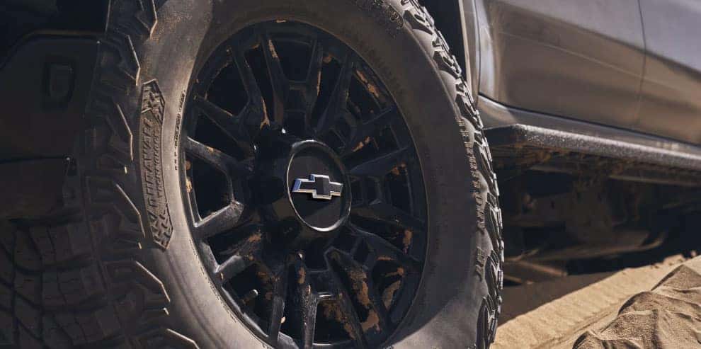 Close-up of a black off-road vehicle tire with a Chevrolet logo in the center. The tire has an aggressive tread pattern, suggesting it is designed for rugged terrain. The vehicles dark metallic body is partially visible in the background.