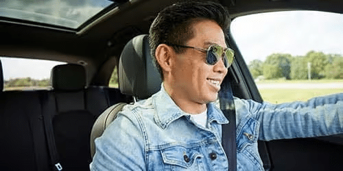 A man wearing sunglasses and a denim jacket smiles while driving a car on a sunny day. The interior is modern, and greenery is visible outside the window.