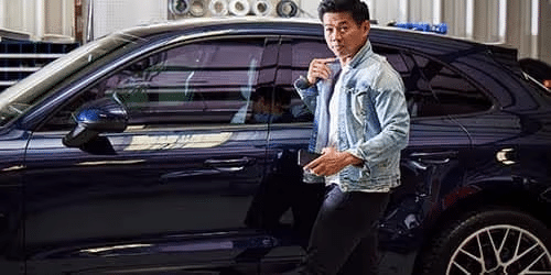 A man wearing a light denim jacket stands next to a dark-colored car in a garage, looking toward the camera. The car’s door is closed and various equipment is visible in the background.
