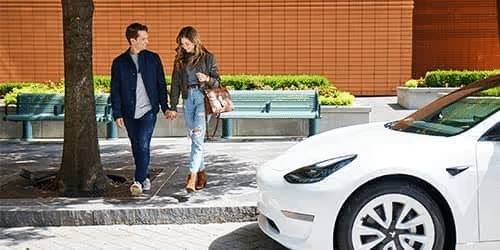 A couple holding hands walks on a city sidewalk near a white car, with benches, greenery, and a brick wall in the background.