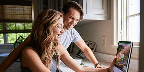 A smiling couple stands in a bright kitchen, looking at a laptop together on the counter near a window, appearing happy and engaged with what they see on the screen.