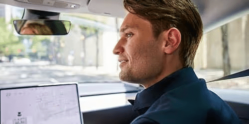 A man with light brown hair sits in the drivers seat of a car, looking forward and smiling, with a navigation screen visible in front of him and a city street scene outside the window.