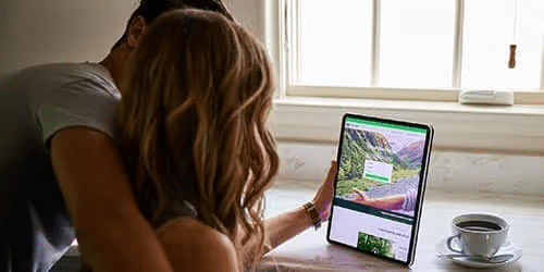 A couple sits together at a table, looking at a tablet with a website on the screen. A window lets in natural light, and a cup of coffee sits nearby.