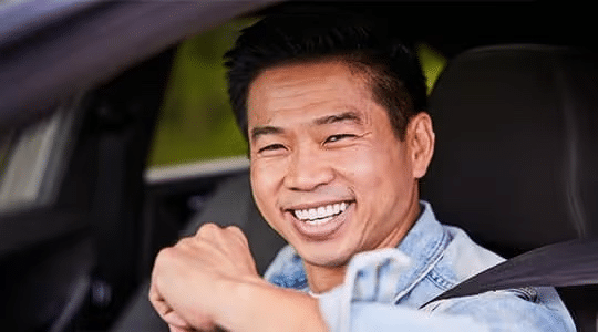 A smiling man sits in the drivers seat of a car, wearing a seatbelt and looking out the open window. He appears happy and relaxed, dressed in a light denim jacket.