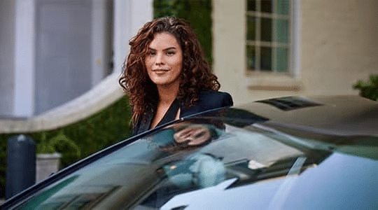 A woman with curly brown hair, wearing a black jacket, stands partially behind an open car door, smiling, with a building and greenery in the background.