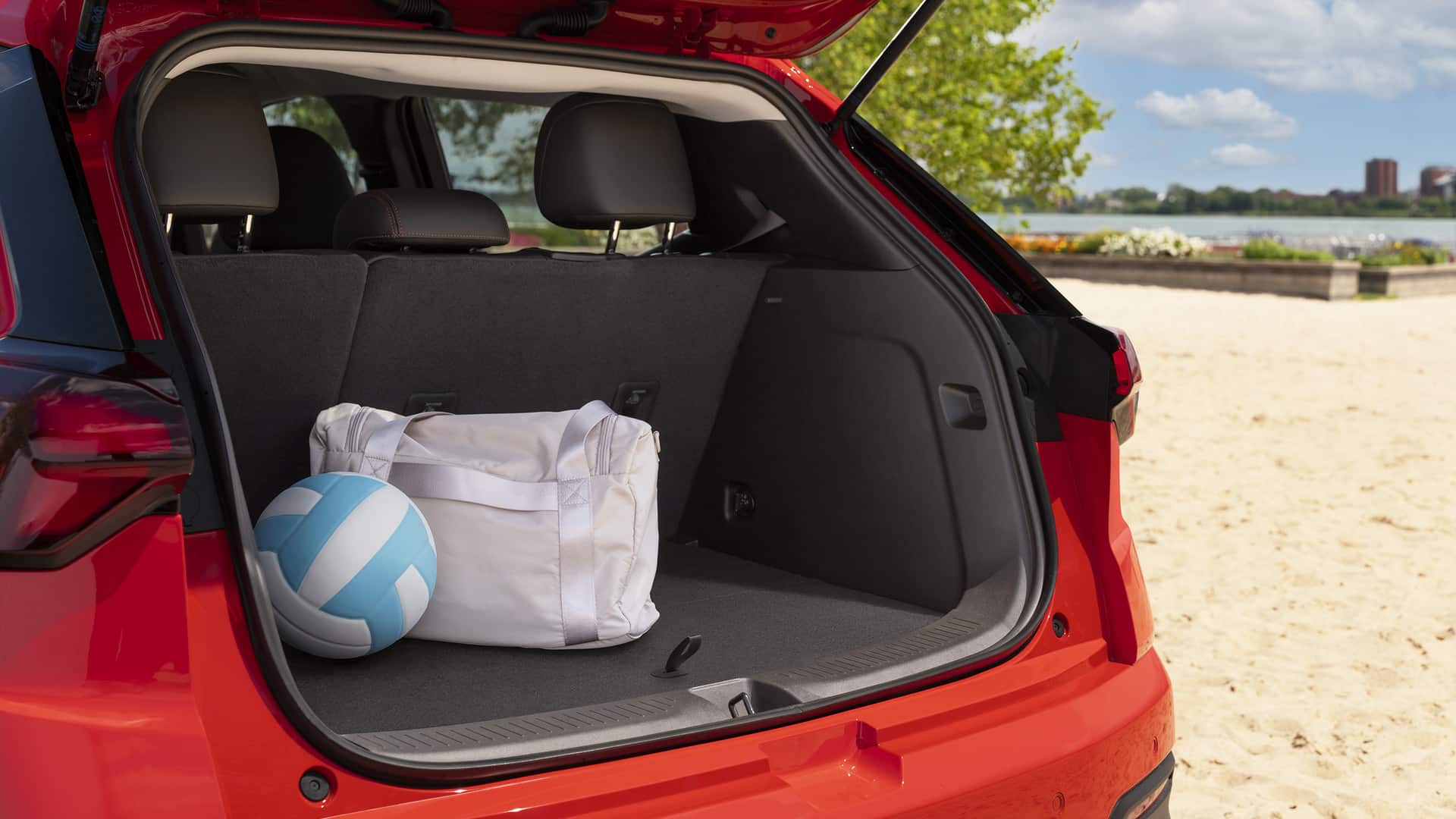 Red SUV trunk with a volleyball and white bag on a sandy beach background.