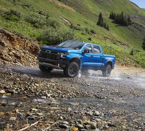 Blue pickup truck driving through a rocky stream in a lush green mountain landscape.