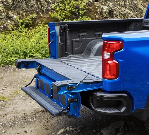 Blue pickup truck with tailgate and step ladder open, parked on rocky terrain and greenery in the background.