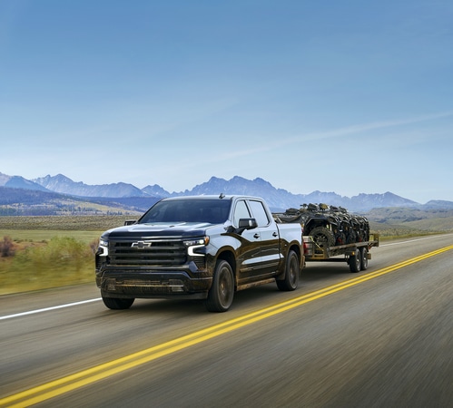 A black pickup truck towing a trailer loaded with motorcycles on a highway, with mountains in the background.