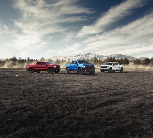 Three pickup trucks in red, blue, and white parked on rugged terrain with a mountain backdrop.