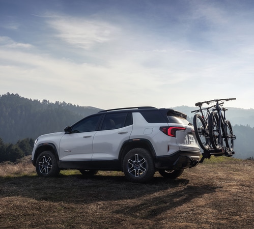 White SUV parked on grassy hill with mountain bikes mounted on the rear, under a clear sky.