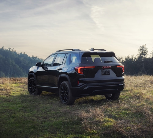 Black GMC SUV parked on grassy hill with a forested background under a cloudy sky.