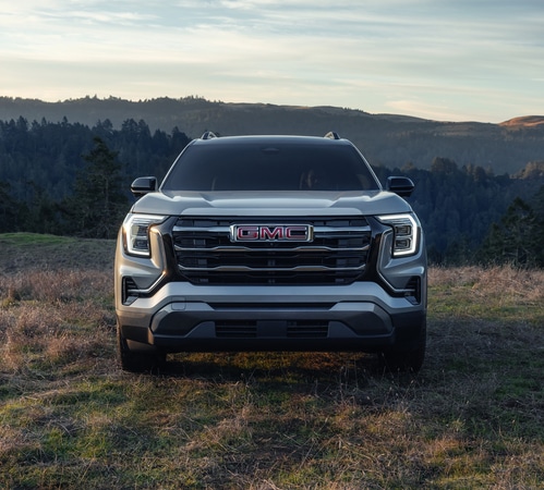 Front view of a modern GMC SUV parked on grass against a forested mountain backdrop.