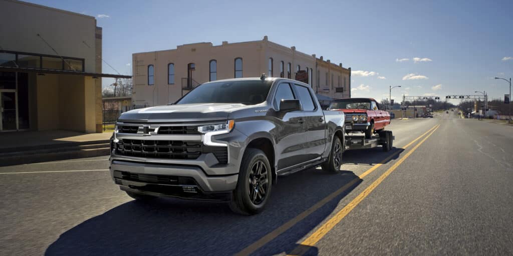 A silver pickup truck towing a red classic car on an empty city street under a clear blue sky.