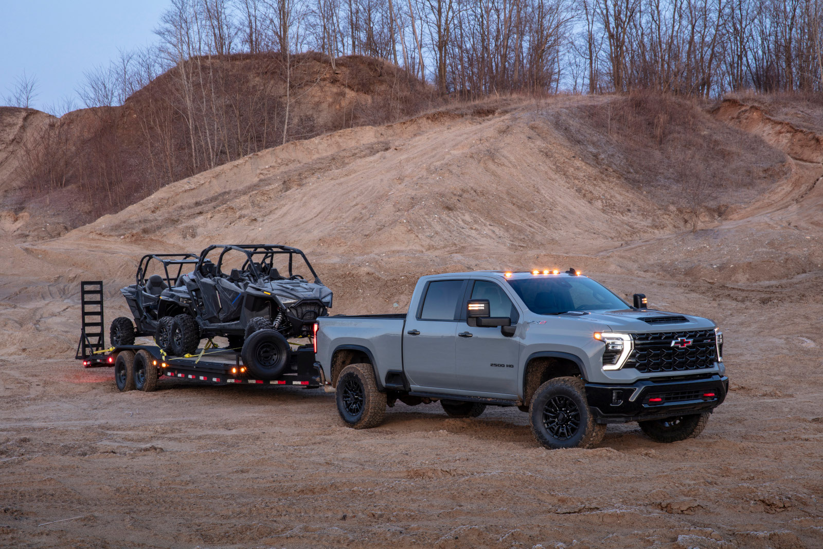 Silver pickup truck towing off-road vehicles on a trailer in a sandy, wooded area.