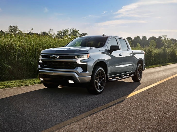 A silver pickup truck driving on a rural road with trees and grass in the background under a blue sky.