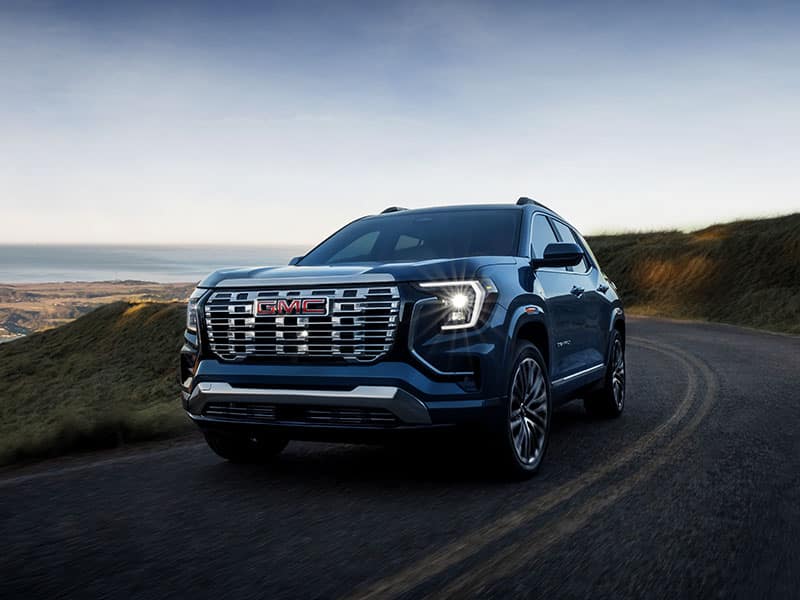 Blue GMC SUV driving on a winding road with a coastal landscape at sunset.