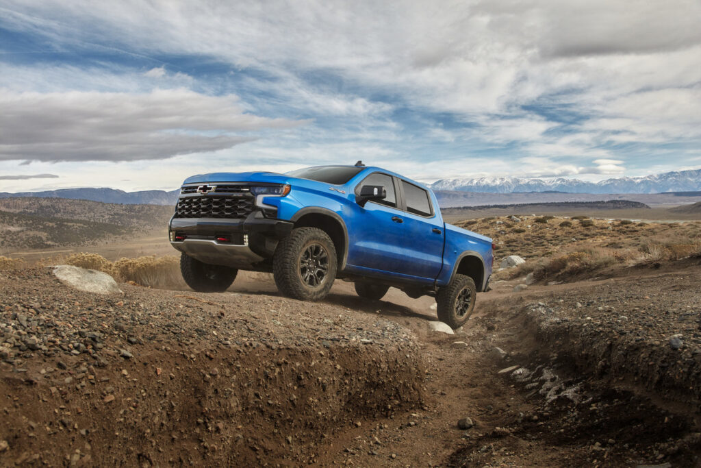Blue pickup truck driving off-road in a rugged desert landscape under a partly cloudy sky.