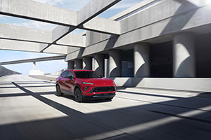 Red SUV driving under modern concrete overpass with blue sky backdrop.
